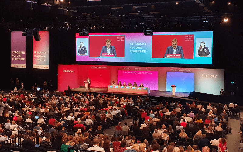 A conference hall of delegates listen to a speaker on the stage