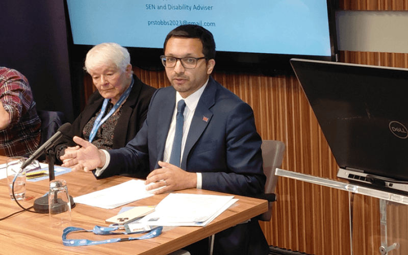 Saqib Bhatti speaking from behind a table, with a woman sitting next to him listening