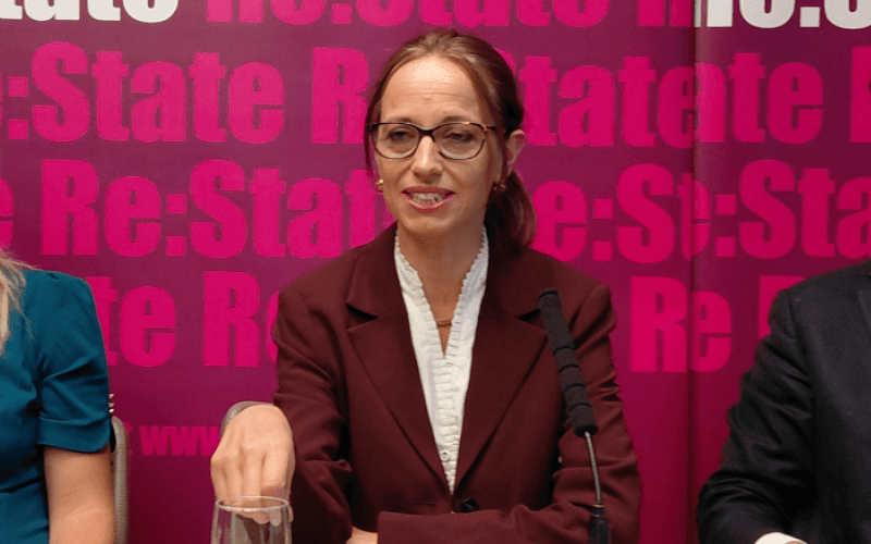 Helen Whately sitting behind a table and a microphone at an event, with the words Re: State behind her on a board