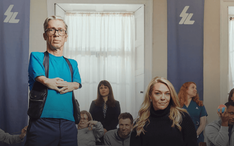Mat Fraser standing and Sophie Morgan in her wheelchair in front of a background of staff and residents in a large room, with a suspiciously SS-type logo on the wall