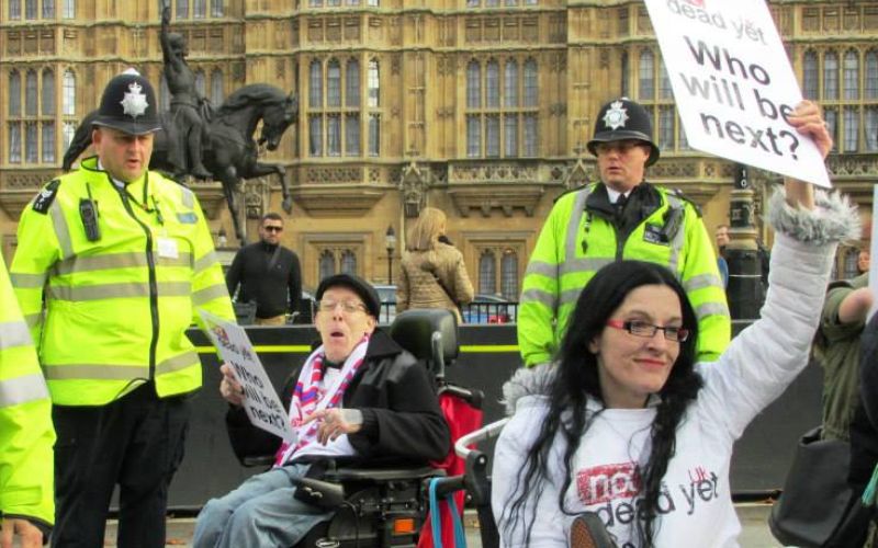 Two wheelchair-users protesting outside parliament, one of them (Dennis Queen) is holding a placard saying 'who will be next', and is wearing a Not Dead Yet UK tee-shirt