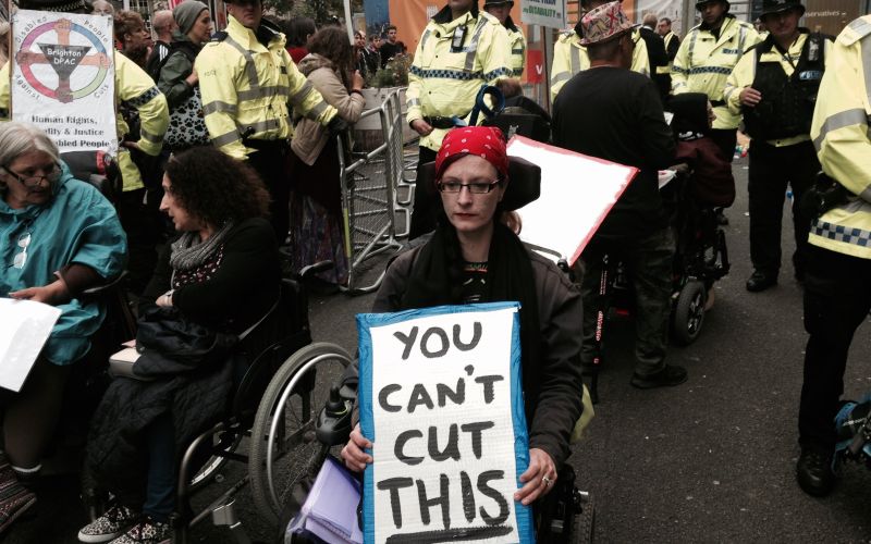 A wheelchair-user at a protest holding a placard saying 'You Can't Cut This'