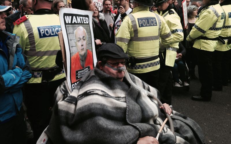 A wheelchair-user wearing a ventilator, with an Iain Duncan Smith 'Wanted' poster behind his head and surrounded by police