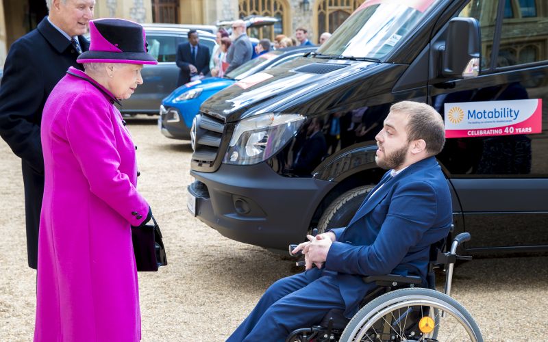 The Queen talks to a bearded man in a wheelchair, with Motability vehicles behind them