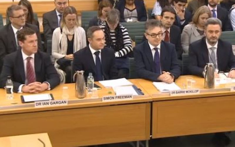 Four executives from Capita and Atos sit behind a table, with other people sat behind them