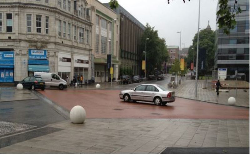 Cars crossing a flat space, with bollards and surrounding buildings