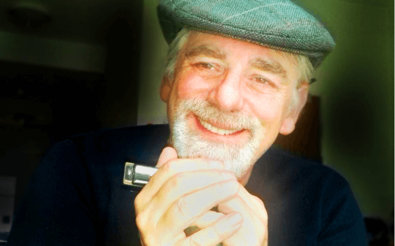 A smiling man in a flat cap holding a harmonica