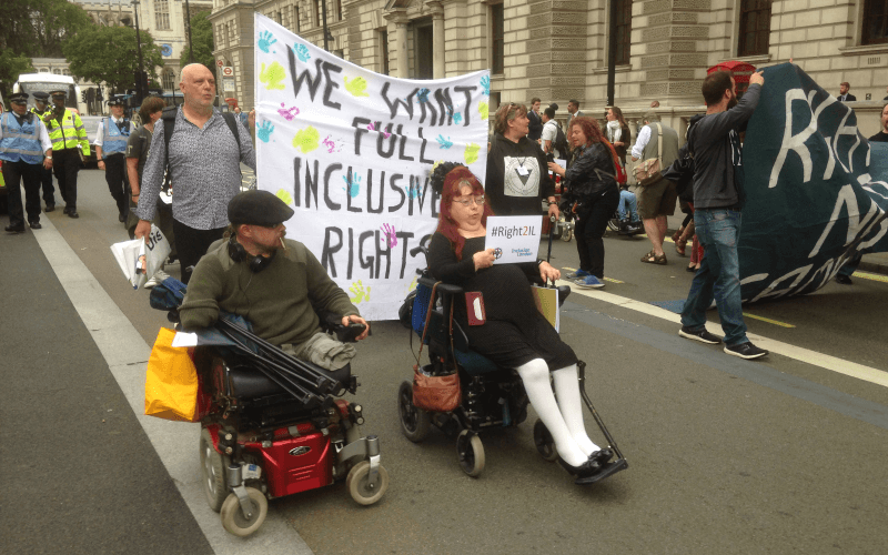 Andy Greene and Penny Pepper in electric wheelchairs at the head of a march in front of a banner calling for full inclusive rights