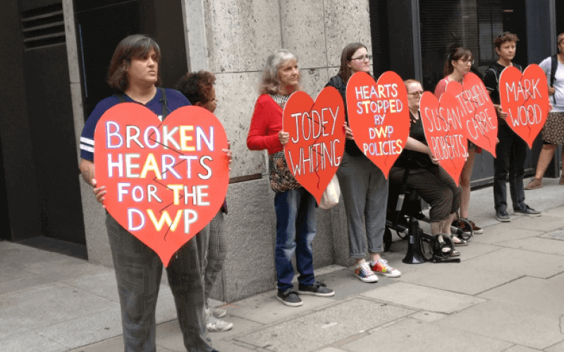 Six women hold large broken red hearts. One has 'broken hearts for the DWP' written on it. Others have names on them.
