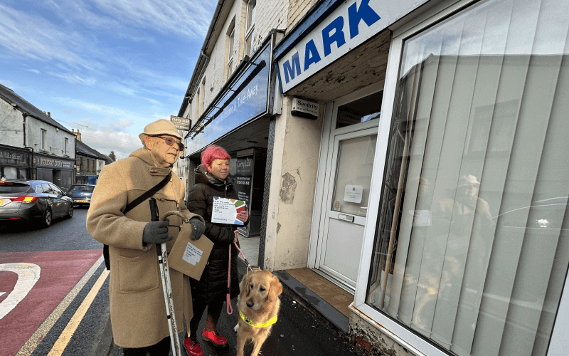 Bill Waddell with a white cane and walking stick, and Sarah Leadbetter and her golden retriever guide dog, with two envelopes outside Mark Harper's constituency office