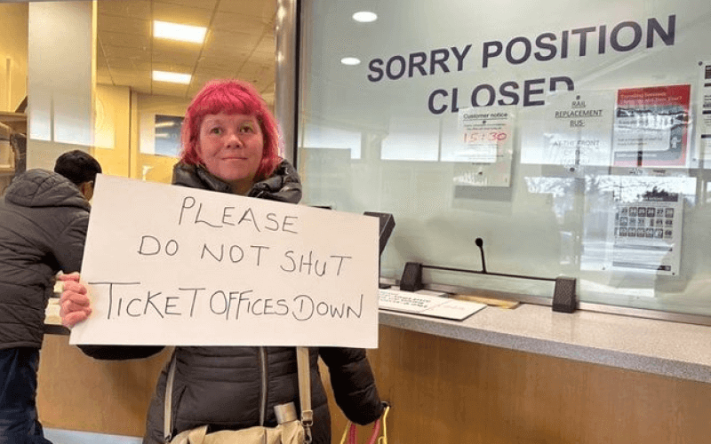 A woman in front of a ticket office which says 'sorry position closed', as she holds a sign saying 'please do not shut ticket offices down'