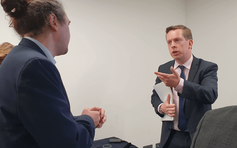 One man listens as another man in a suit, holding some papers, speaks to him across a table