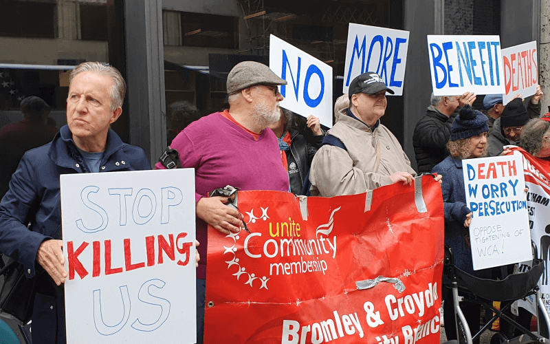 People holding banners outside a building, with messages such as 'Unite Community', 'No more benefit deaths', 'stop killing us', and 'death worry persecution, oppose tightening of WCA'