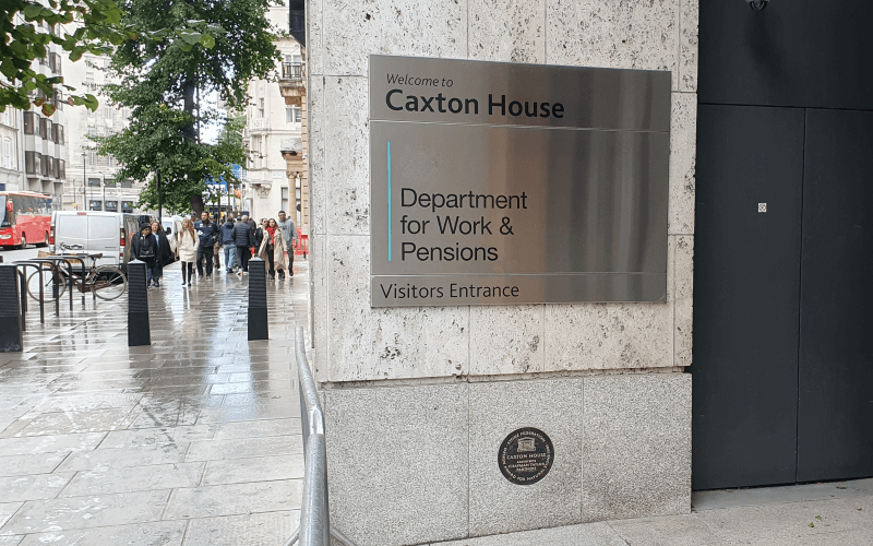 A silver sign on a wall says Welcome to Caxton House, Department for Work and Pensions, Visitors Entrance, with people walking towards the camera on the pavement to the left