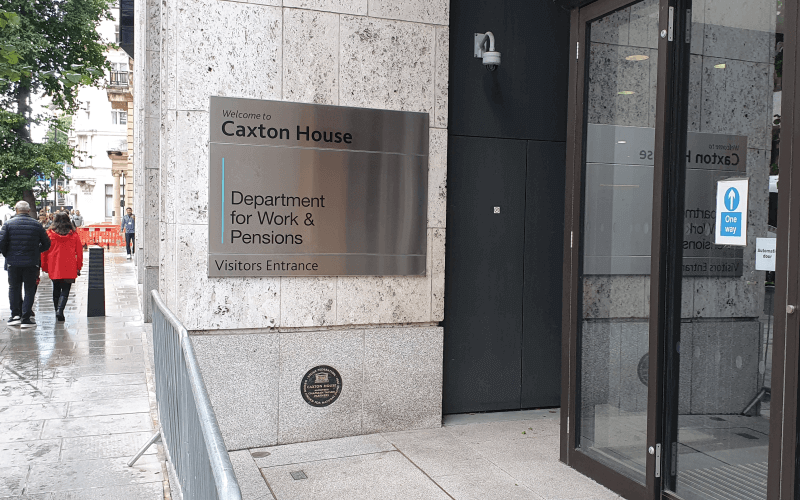 A silver sign on a wall says Welcome to Caxton House, Department for Work and Pensions, Visitors Entrance, with people walking away from the camera on the pavement to the left