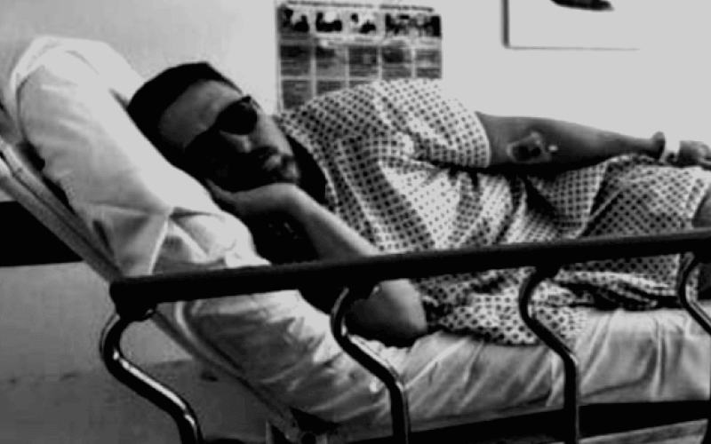 A man wearing dark glasses, lying on his side in a hospital bed, black and white picture