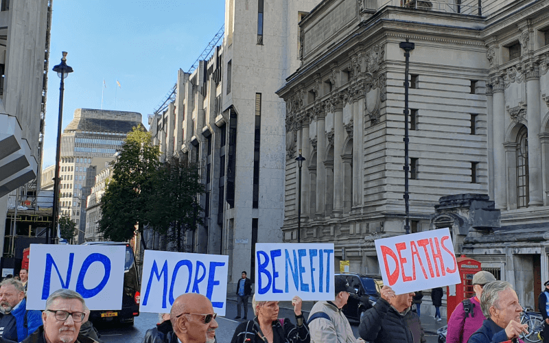 Protesters blocking a street, with the DWP's offices in the background, hold up placards saying 'no more benefit deaths'