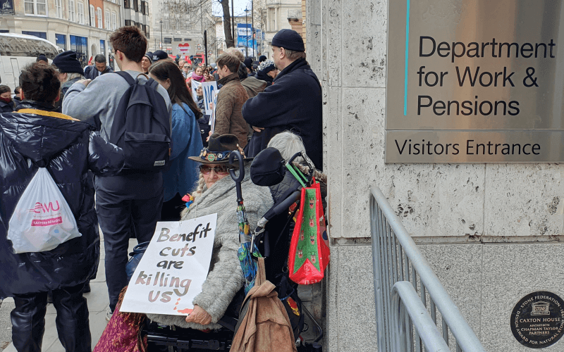 A women sits in her wheelchair holding a sign that says 'Benefit cuts are killing us', with protesters stood behind her, as she sits next to a silver sign on a wall that says Department for Work and Pensions