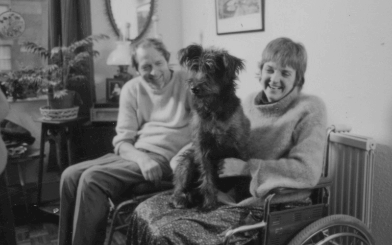 Black and white photo of a woman in a wheelchair with a cute, scruffy dog on her lap, and a man sitting behind them; both are laughing.