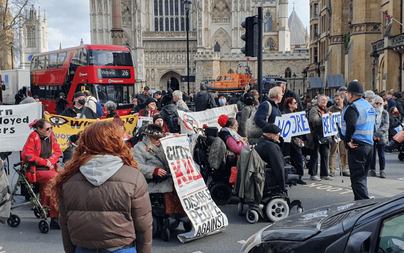 Protesters, including several in wheelchairs, block traffic in front of Westminster Abbey, while holding placards. One placard says 'Cuts Kill'