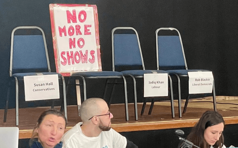 Three people on a panel in front of a stage on which there are four chairs, marked Susan Hall, Sadiq Khan and Rob Blackie, with a sign saying 'No More No Shows' on the fourth chair