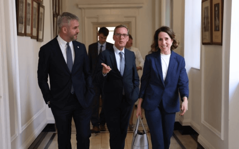 Two men and a woman, all wearing dark suits, walk down a corridor towards the camera