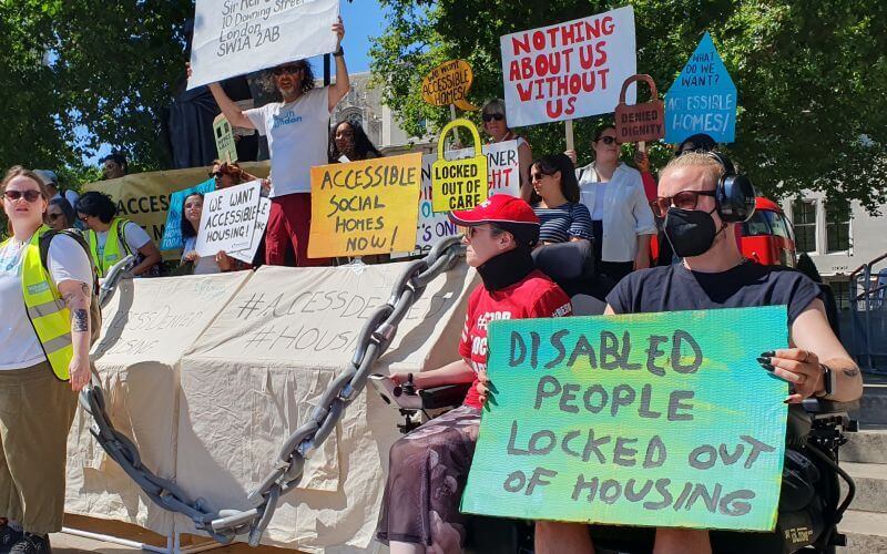Protesters, some in wheelchairs, in front of a statue of Mahatma Gandhi, holding placards calling for action on accessible housing, including 'disabled people locked out of housing', beside a big silver chain.