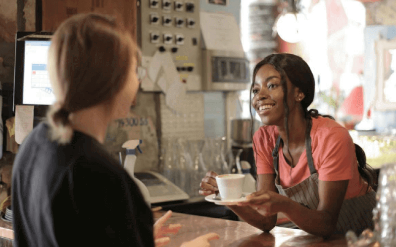 A smiling young woman in an apron leans over a counter in a coffee shop to hand a cup of coffee to a customer
