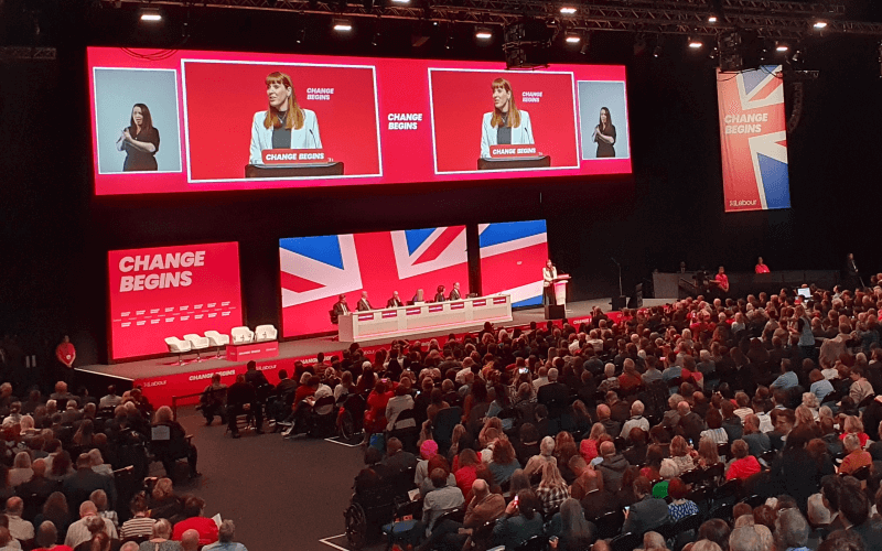 Delegates watch Angela Rayner on stage and on a big screen behind her, with a large union flag and the message 'change begins' behind her