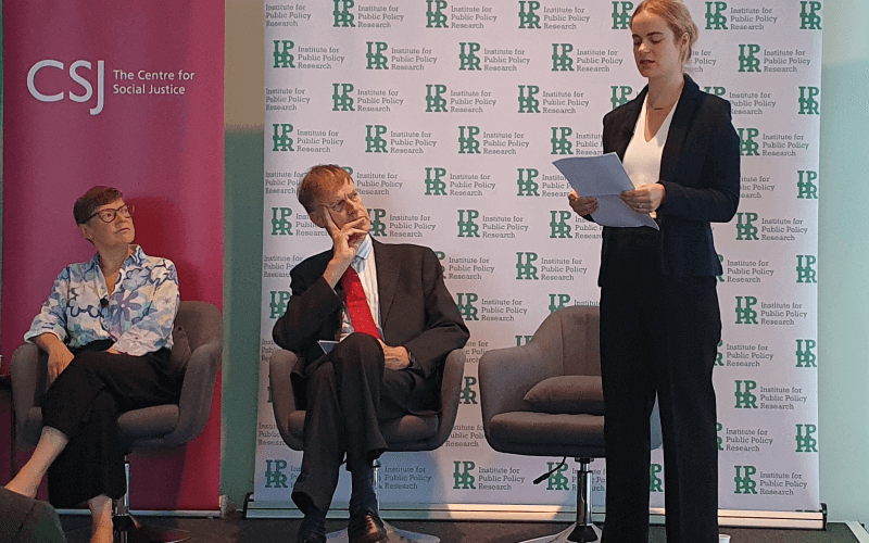 Sir Stephen Timms sits and watches a woman delivering a speech, with another woman sitting next to him on the stage
