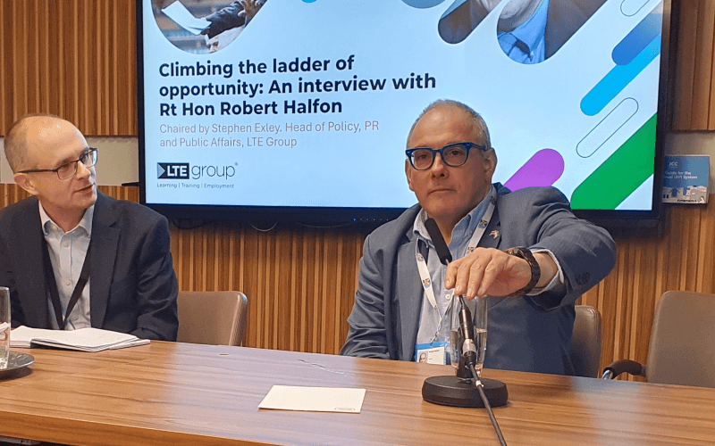 Robert Halfon adjusting a microphone on a table, with another man sat next to him asking him a question. Behind them a screen shows the words Climbing the ladder of opportunity: an interview with Robert Halfon.