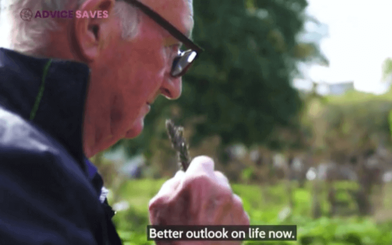 A man sniffs a plant, as he looks pensive, with an allotment in the background