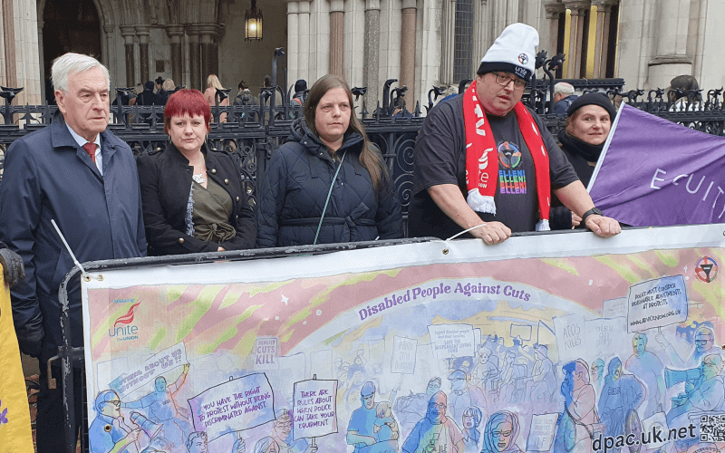 John McDonnell, Ellen Clifford and other activists stand in front of the Royal Courts of Justice and behind a long painted banner for Disabled People Against Cuts, which features many disabled protesters at various actions