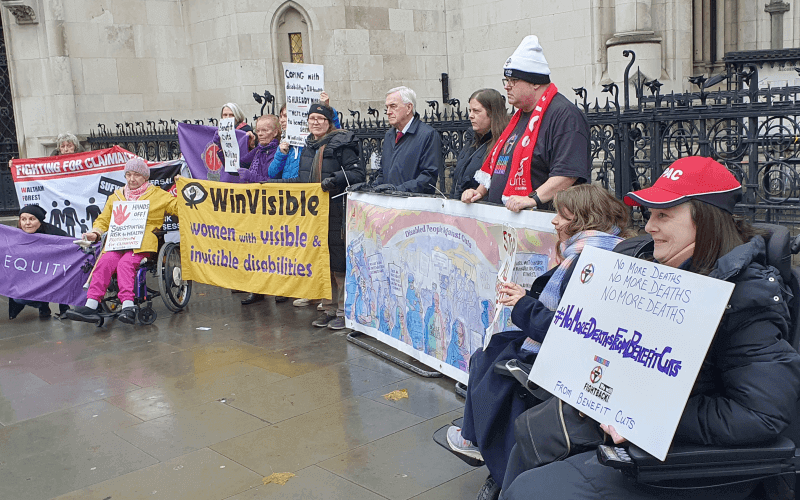 Activists in front of the Royal Courts of Justice, with banners and placards, including a long painted banner for Disabled People Against Cuts that shows people at various protests, and in the foreground a woman in a red cap holds a placard saying 'No more deaths from benefit cuts'