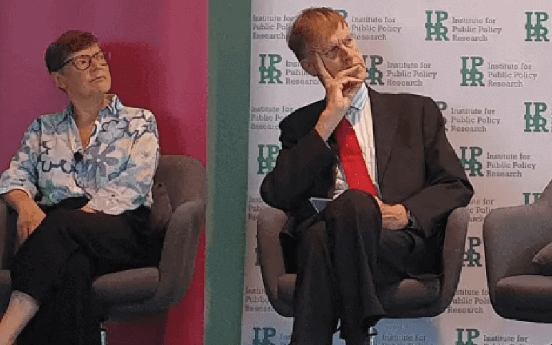 Sir Stephen Timms sitting on a stage with his head resting in his right hand as he listen to someone on his left, with a woman sitting on his right, and multiple logos for IPPR on a screen behind them