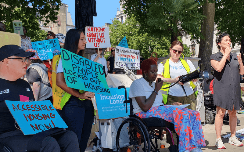A black woman delivers a speech from her wheelchair, in front of a statue of Mahatma Gandhi and protesters holding placards calling for action on accessible housing, including 'disabled people locked out of housing' 'nothing about us without us' and 'accessible housing now'.