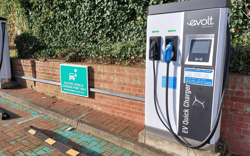 An electric vehicle charge point in front of a brick wall and placed on a kerb, with a hedge behind