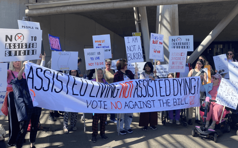 Disabled protesters and allies holding multiple placards opposing assisted suicide, and a long banner, with messages including assist us to live, not die; I am not a burden; and vote no against the bill