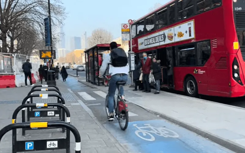 Two passengers, one of them wearing a mask, wait to cross a cycle lane onto a floating bus stop after leaving a bus, while a cyclist rides along the cycle lane